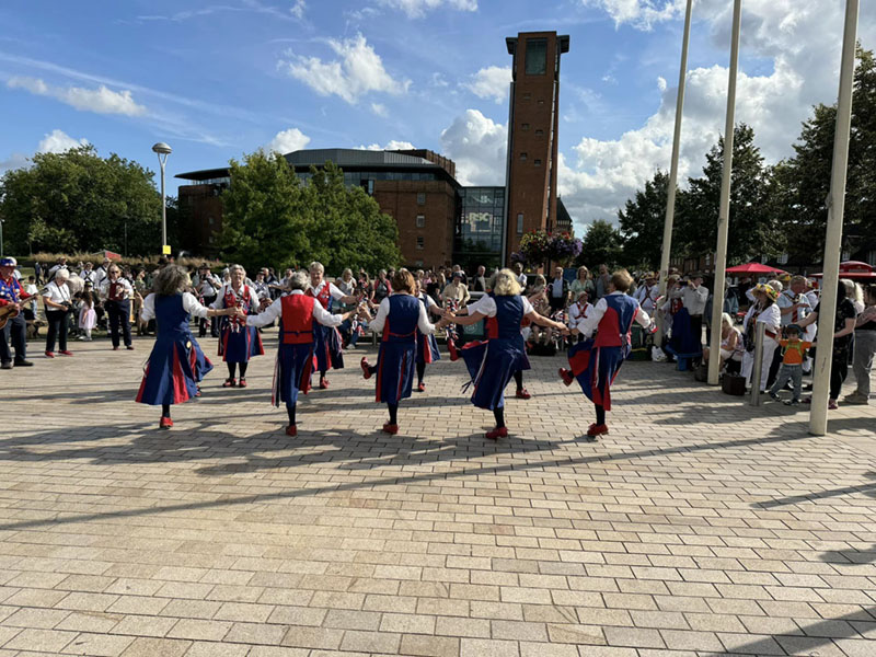 8 dancers in 2 neat rows of 4 dancing in the gardens in Stratford, with the Shakespeare theatre in the background, against a blue sky.