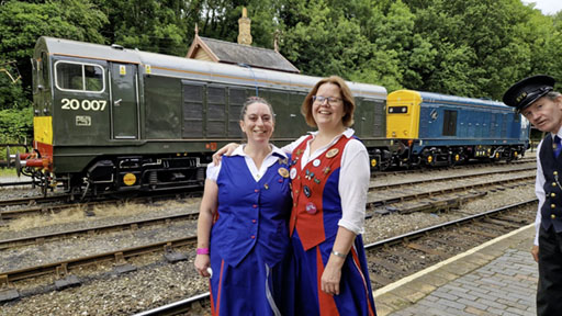 two dancers standing on a platform with two large diesel engines in the background. A uniformed member of station staff looks on.