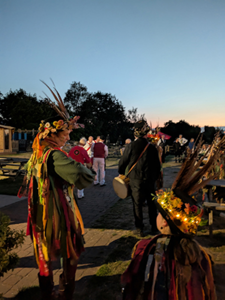 2 of Bow Brook Border Morris dancers with illuminted head wear as the night draws in, sitting in front of White Hart Morris dancers in action