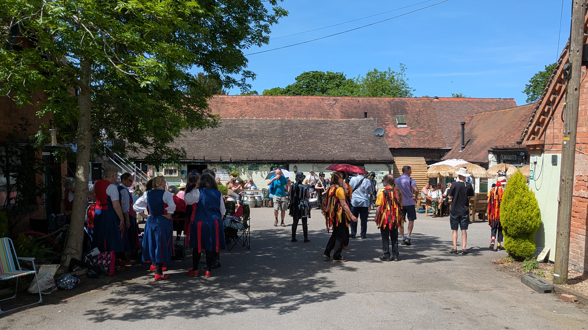 general view of the site showing sunlit dancers preparing to perform, others under shade of trees, and several spectators sitting ready to watch the next performance